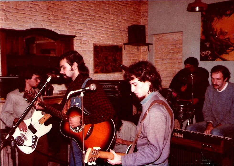 Un grupo de músicos jóvenes tocando guitarras, bajo, batería y teclado en una sala con paredes de ladrillo